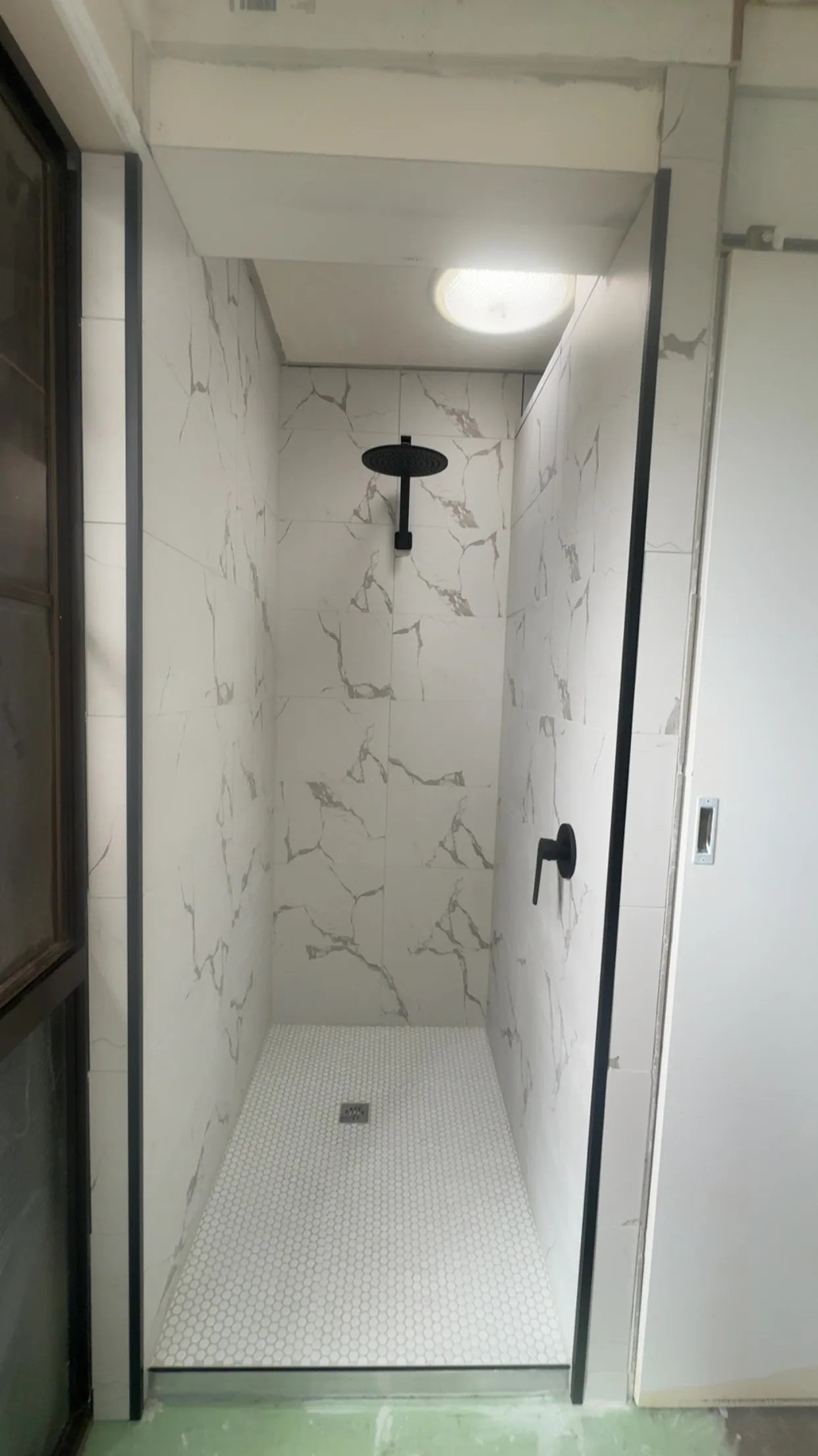 After: newly tiled walk-in shower recess with large-format marble-look wall tiles, white hexagonal floor tiles, and matte black shower rose and fixtures
