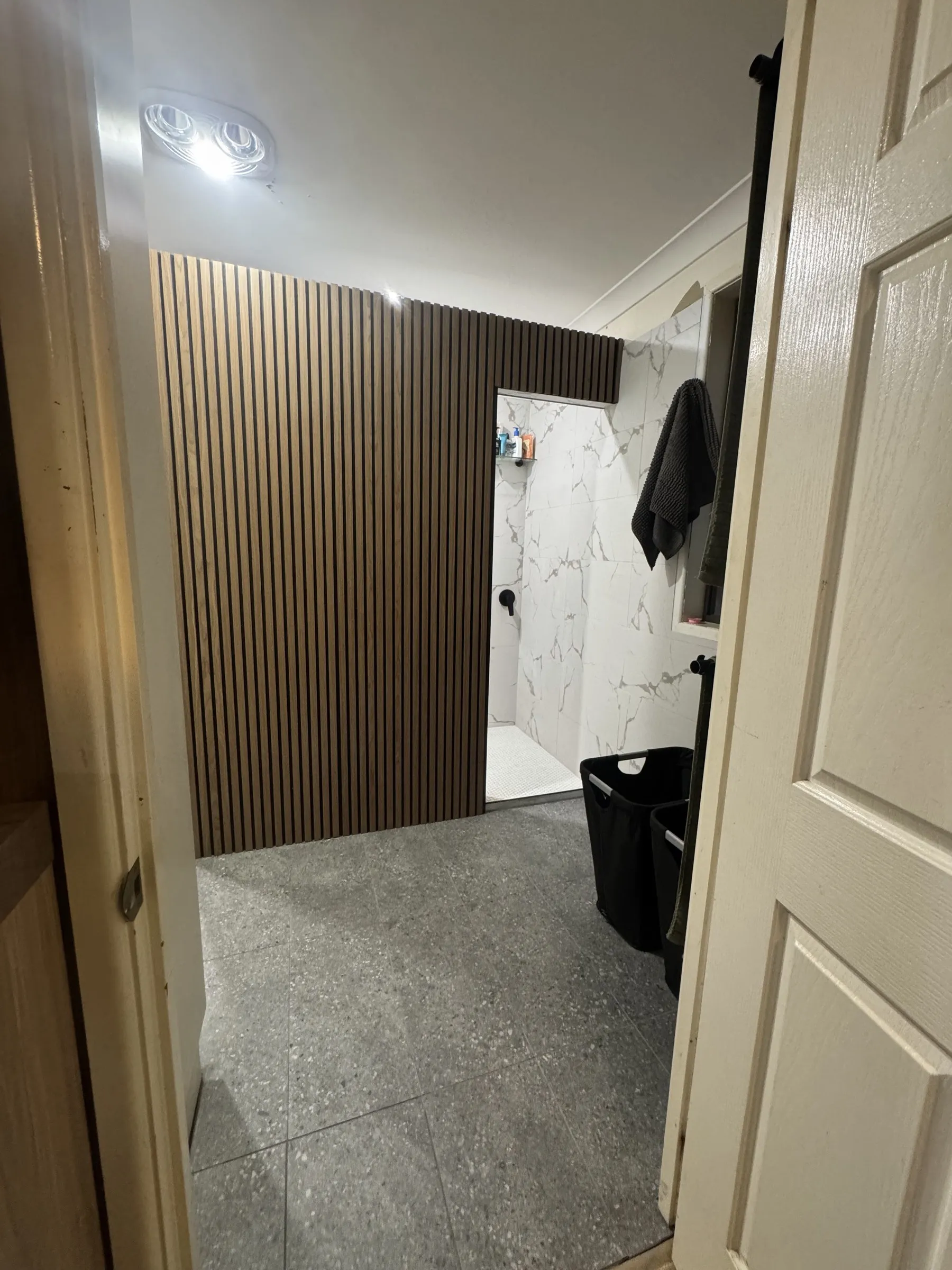 After: completed bathroom renovation showing timber slat feature wall, large-format grey floor tiles, and marble-look wall tiles glimpsed through the doorway