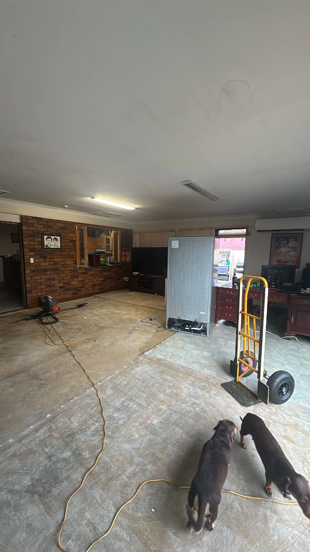 Before: large open living or garage space with concrete floor and brick feature wall, with existing belongings in place prior to the renovation works