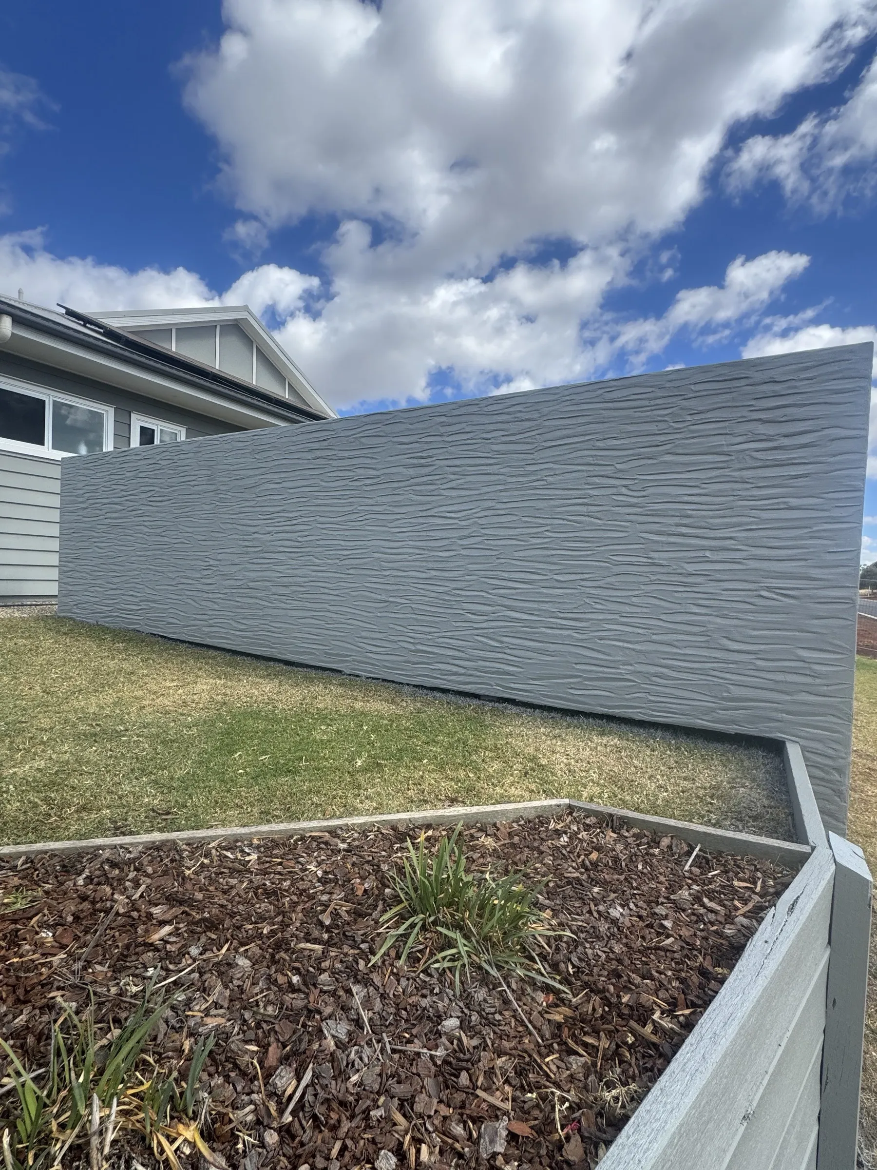 Close-up of a textured grey panel custom fence beside a garden bed, showing the wave-relief surface finish and clean install alongside a modern home