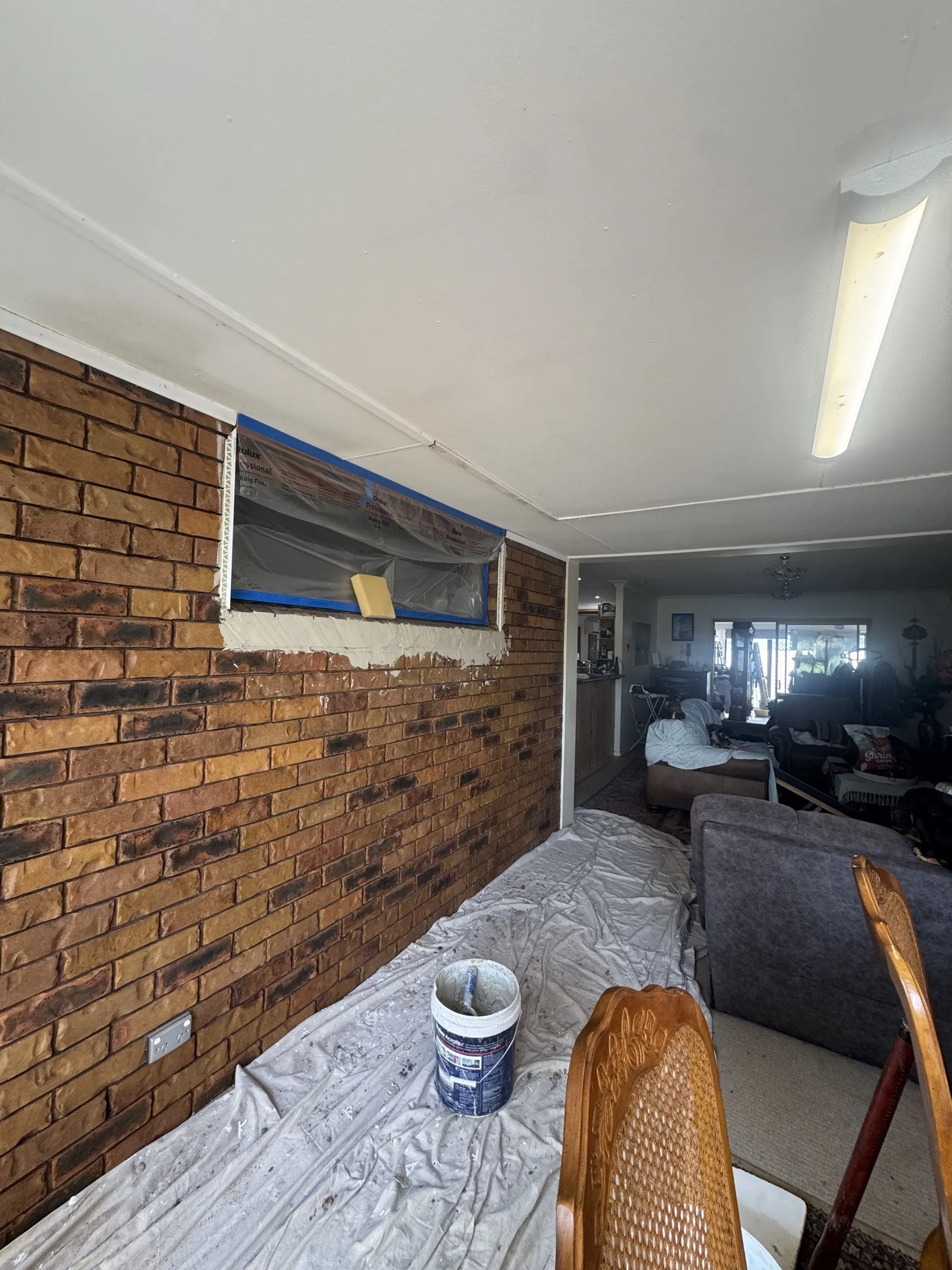 Before: interior of a home showing original exposed brick wall being prepared for painting, with drop sheets protecting furnishings during the job