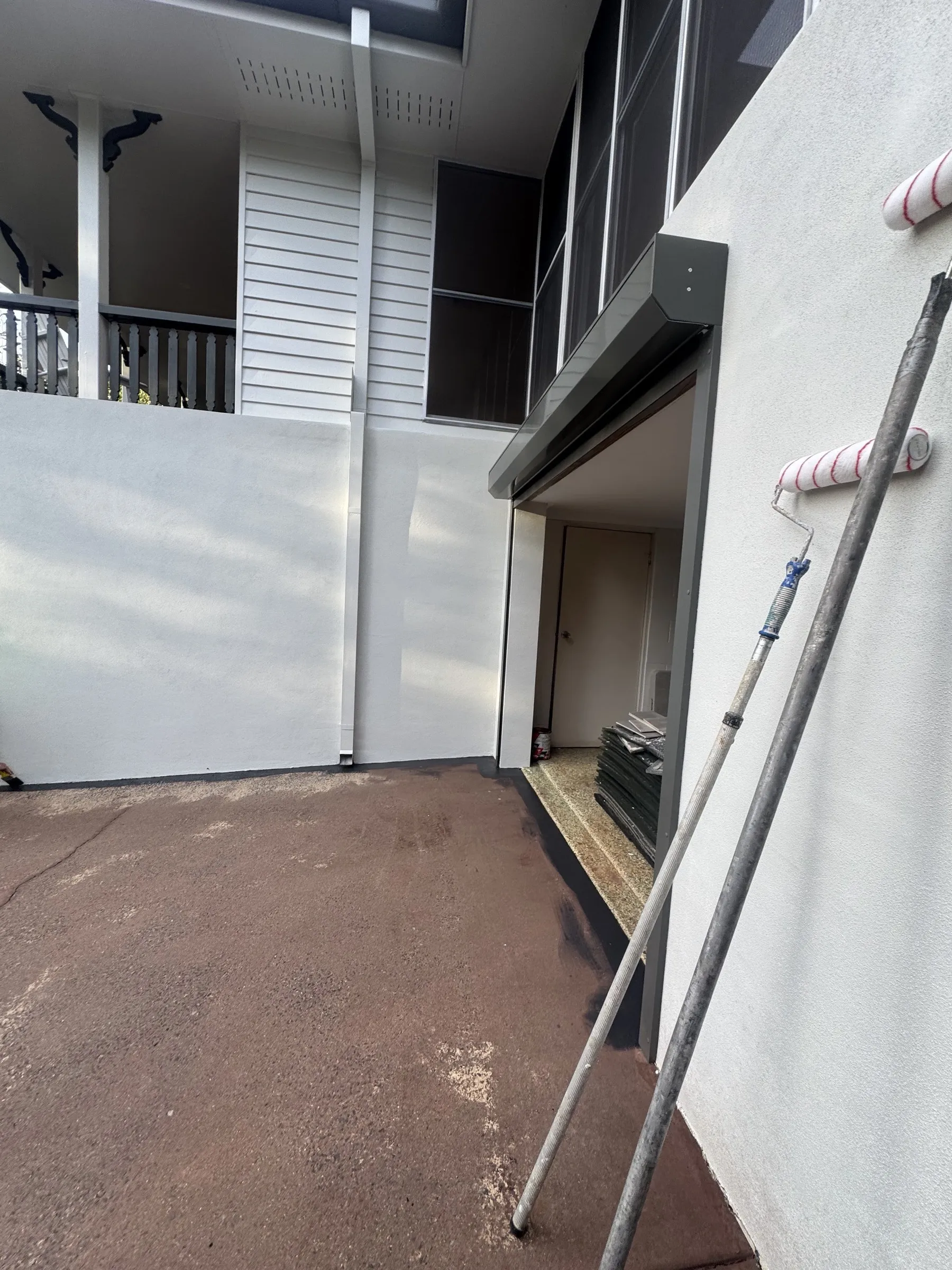 Exterior painting in progress on a two-storey home — roller poles and paint equipment leaning against freshly rendered white walls, with dark trim and balcony above
