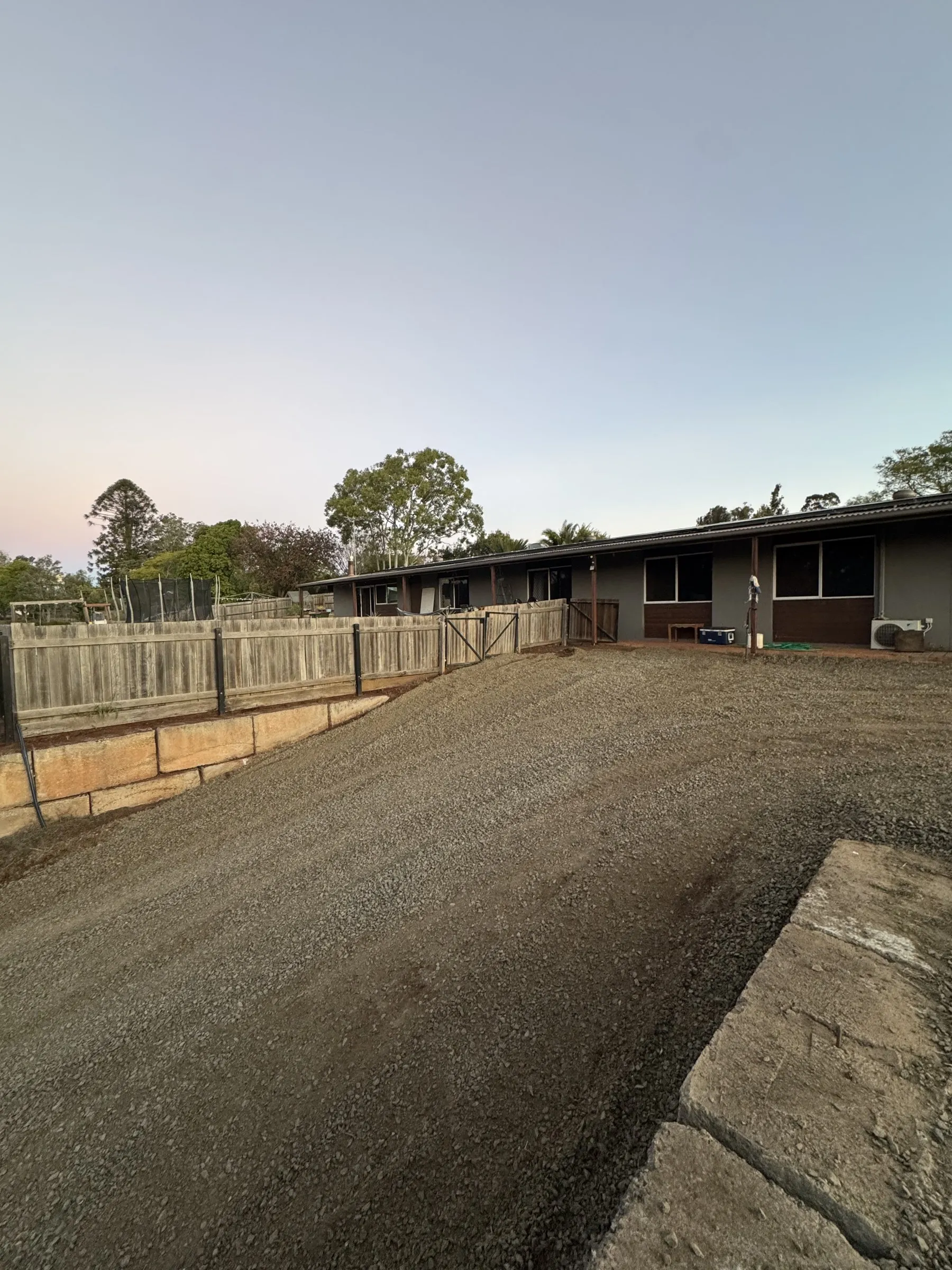 A long timber sleeper retaining wall holding a freshly graded earth pad beside a rural home at dusk, part of a full yard transformation in the Southern Downs